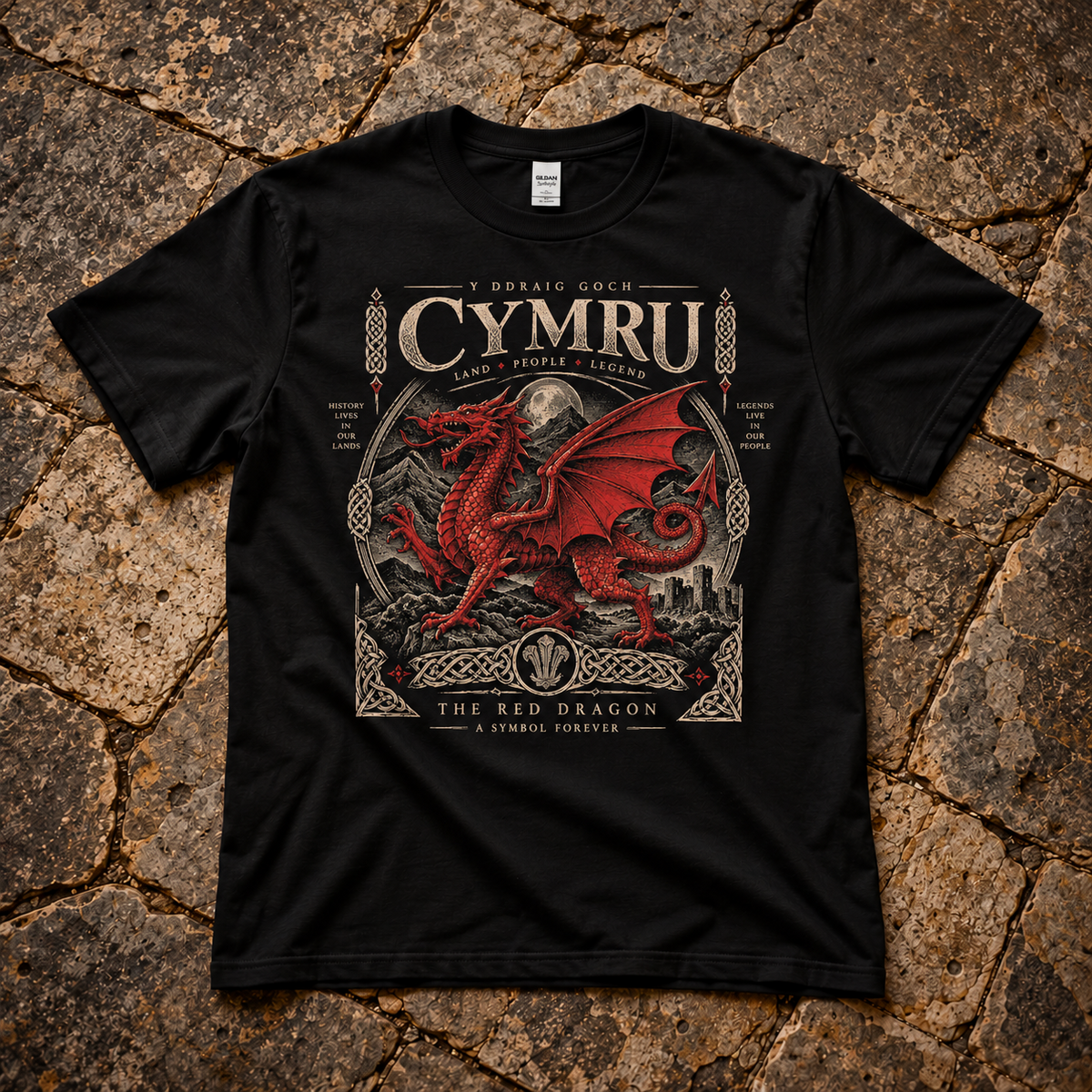 A black short-sleeve graphic T-shirt laid flat on a textured stone surface. The shirt features a large front print with the words 'CYMRU', a red dragon illustration, and additional decorative text in Welsh and English.